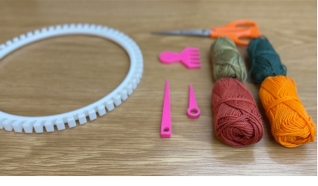 Image of white 3D printed loom, pink 3D printed weaving comb and knitting needles, orang handled scissors, and 4 bundles of yarn in light green, dark green, red, and yellow.