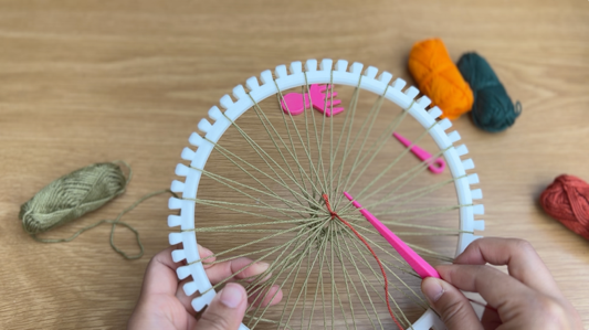 Image of white plastic loom, now fully looped with a green yarn base structure. A pink needle is being used to loop red yarn through the strands of the base structure.