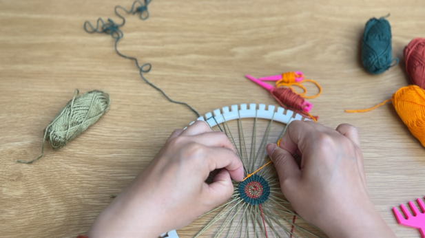 Image of the white plastic loom with a woven green structure and several rows started. Yellow yarn is being tied to the dark green yarn.