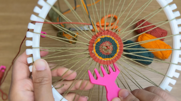 Image of a white plastic loom with a woven base structure. A pink weaving comb is being used to push yarn towards center of the structure.