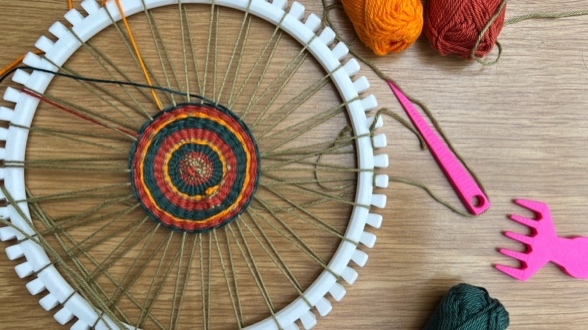 Image of a white plastic loom with a half-finished striped woven circle. Bundles of coloured yarn and pink weaving accessories sit to the side.