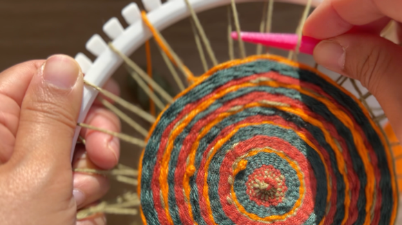 Close-up image of a white plastic loom with a nearly finished, colorful woven circle. A pink needle is being used to weave in another row.
