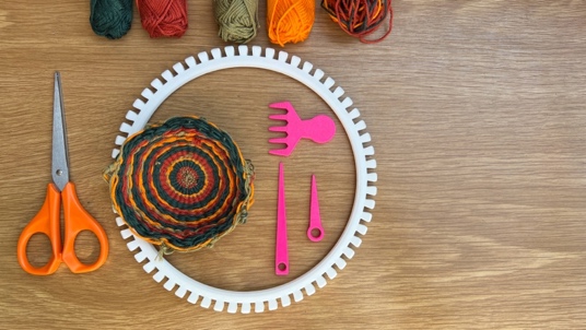 Image of a circular, patterned woven piece, alongside the tools used – including a 3D-printed white miniature loom.