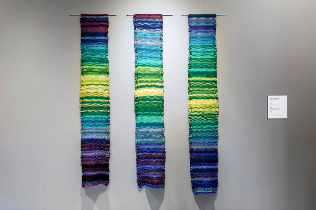 Image of three knitted wall hangings. They are striped in shades of green, blue, and pruple.
